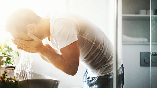 Morning bathroom refreshment. Closeup side view of partially unrecognizable man splashing his face with water over bathroom sink. Brightly back lit with sunlight. He's wearing pajamas and white t-shirt. vanity mirror stock pictures, royalty-free photos & images