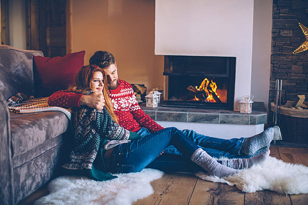 Couple on vacation at mountain cabin. Sitting on the floor on a fur by fireplace in cozy  living room on Christmas. Wearing festive knitted sweaters and socks. Austrian Alps.