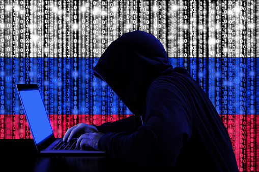 Hacker in a dark hoody sitting in front of a notebook with digital russian flag and binary streams background cybersecurity concept