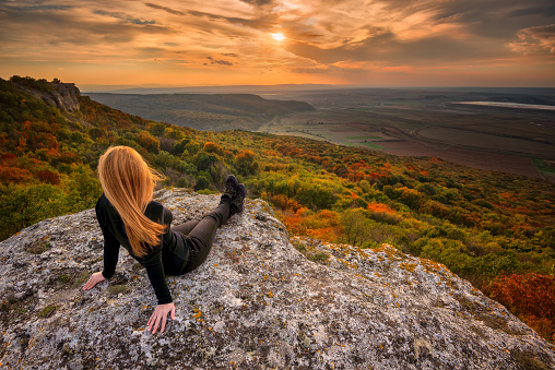 A woman on the top of a rock enjoys the view of sunset over an autumn forest