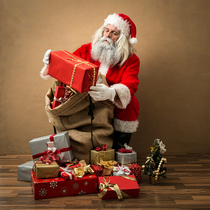 Santa Claus looking at a gift in his hand. There are lots of red and golden presents in his bag. He is sitting in front of a brown wall and de is wearing the traditional clothing with the red cap, jacket and trousers, the black belt and golden glasses. The photo was shot in the studio.