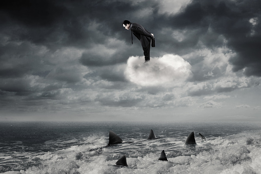 Young businessman standing on the cloud while looking down at dangerous sharks on the sea