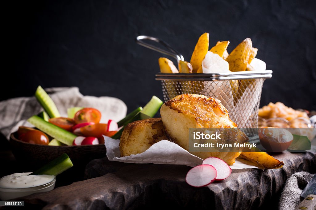 Fried fish fillet with baked potatoes Fish and chips. Fried fish fillet with homemade baked potatoes and fresh salad on wooden cutting board. Backgrounds Stock Photo