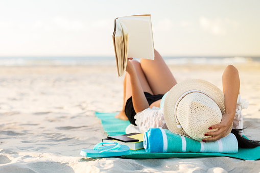 A woman lies on her back on the beach. She is holding up a hard cover book. She is lying on a mat with a rolled up beach towel underneath her head. She is also wearing a straw hat. A pair of flip flops, smart phone and another book are beside her. The sandy beach and ocean are blurred in the background. Copy space is available.