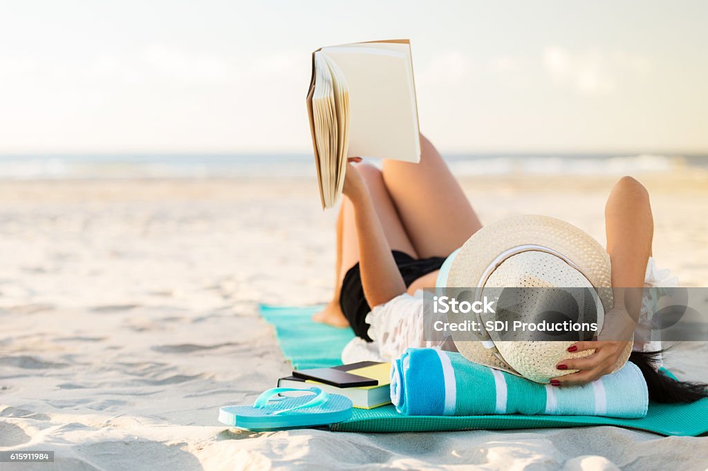 Woman lies on the beach reading a book A woman lies on her back on the beach. She is holding up a hard cover book. She is lying on a mat with a rolled up beach towel underneath her head. She is also wearing a straw hat. A pair of flip flops, smart phone and another book are beside her. The sandy beach and ocean are blurred in the background. Copy space is available. Beach Stock Photo