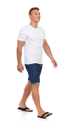 Young man in white shirt, jeans shorts and black sandals walking and looking away. Front side view. Full length studio shot isolated on white.