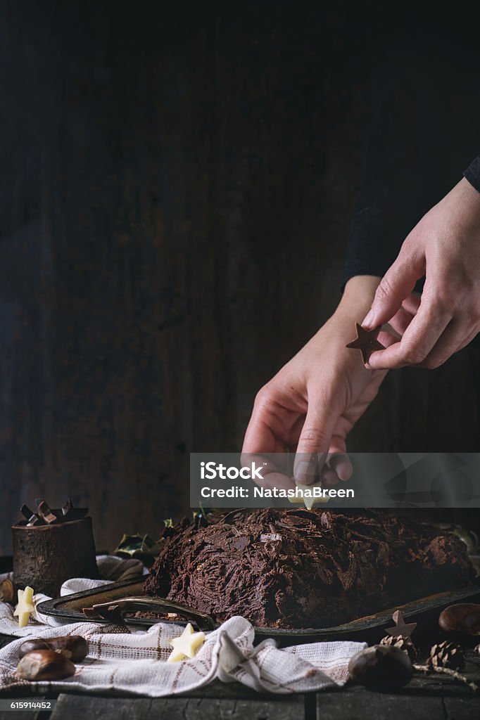 Decorating of christmas chocolate yule log Decorating process of homemade Christmas chocolate yule log by woman's hands with chocolate stars over old wooden table with holly branch and chestnuts. Dark rustic style. With retro filter effect Activity Stock Photo