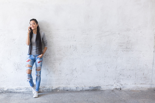 Smiling young asian woman leaning back on blank wall and talking on smart phone. Wears ripped jeans and gray blouse, with long black hair. Looking away, one hand in pocket.
