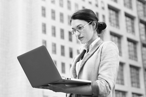 Business woman in front of building, working on a laptop. Holding the laptop in her hands.