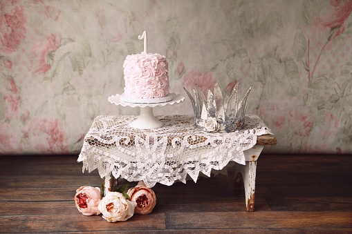 A pretty pink first birthday cake, and a princess crown on a rustic wooden bench. vintage pastel rose wallpaper in background. Cut peonies rest on the hardwood floor. 