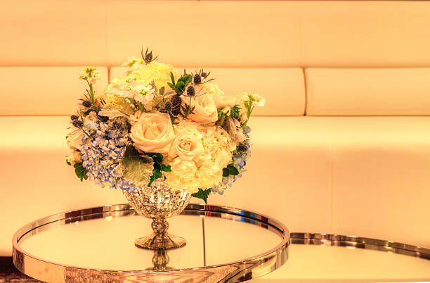 Wedding bouquet in a silver vase on a mirror table with white chairs and a sofa in the background. Flowers include white poppy Papaver species, white roses, blue thistle flowers, hydrangea and sweat pea.