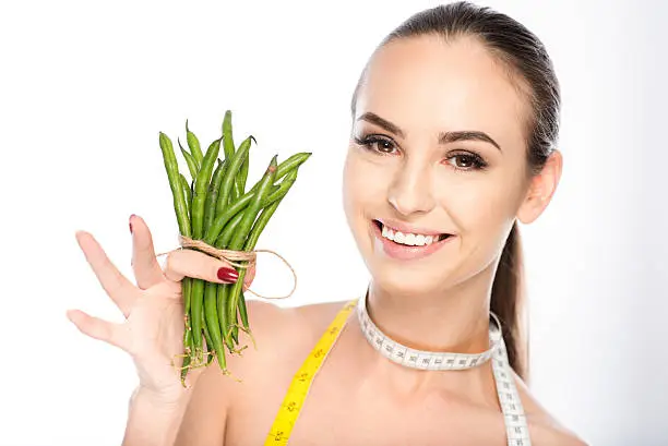 Slim young girl is showing green beans and laughing. She is carrying tape on neck. Isolated
