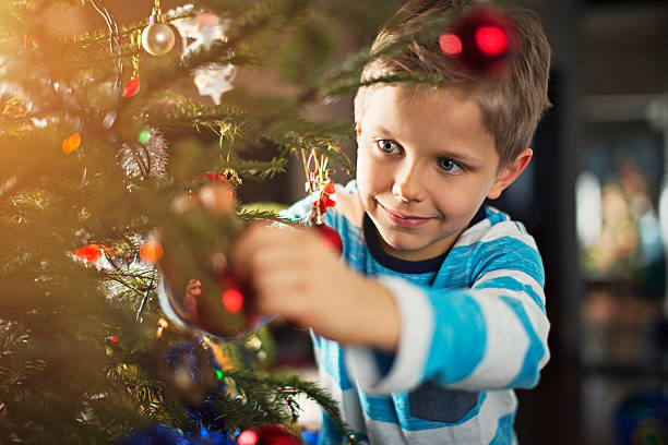 Cute little boy decorating christmas tree. The boy is aged 6 and is wearing casual clothes. The boy is smiling and is hanging a bauble on the christmas tree
