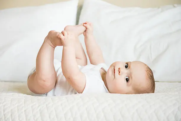 Happy baby boy in white bodysuit holds on to toes while playing, lying on back on bed
