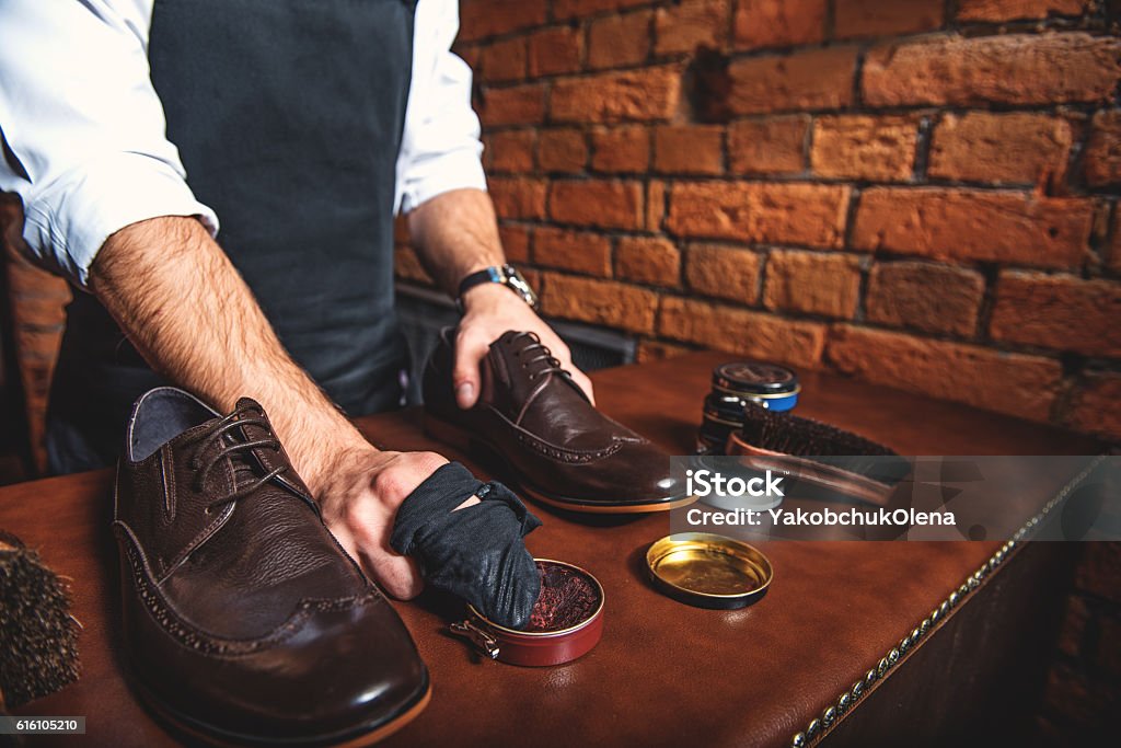 worker polishing a pair of shoes polisher standing at the desk with a boot and rag in his hands Adult Stock Photo