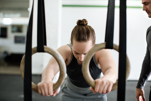 Young woman is doing exercises on gymnastic rings with the help of a personal trainer in a gym