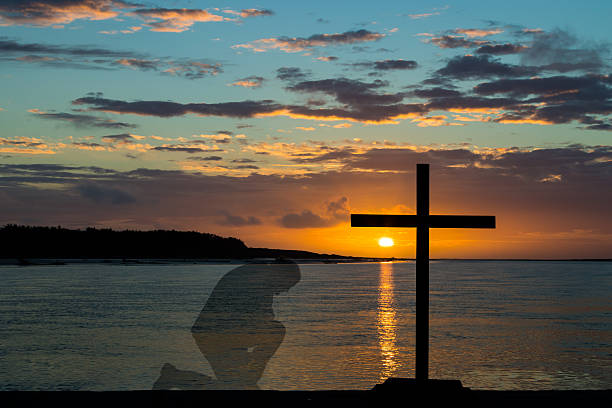 Black cross and a man kneeling, with a sunset at a river mouth area.