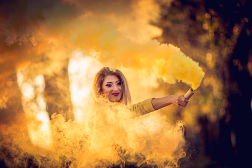 Young pretty girl outdors in a park, on a lovely autumn day, holding a torch, there is yellow smoke all around her