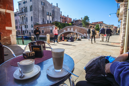 Venice, Italy - October 12, 2016: Tourists sitting in a cafe in Venice, Italy. A canal is passing the scenery and a bridge is crossing the water. Venice is a famous italian city with lots of canals. It is situated on lots of small islands and is famous for the gondolas shipping on the water.