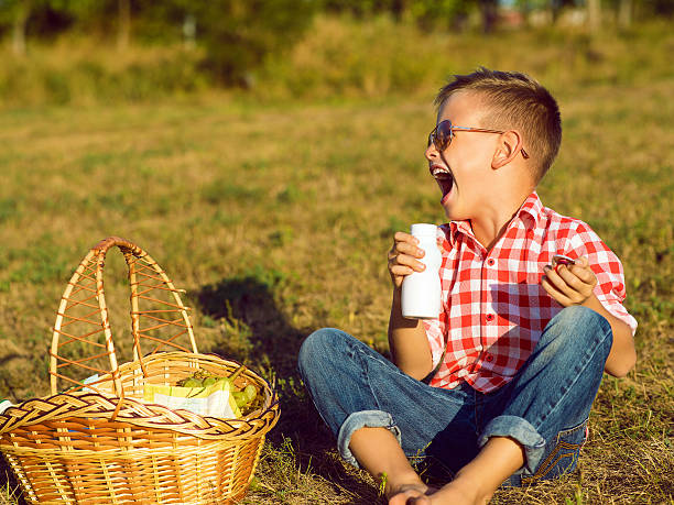 Little stylish kid wearing trendy checked shirt and jeans is having fun on the picnic. He is drinking yogurt and has milk moustache over his mouth. Dairy products concept. Outdoor shot. Copy-space