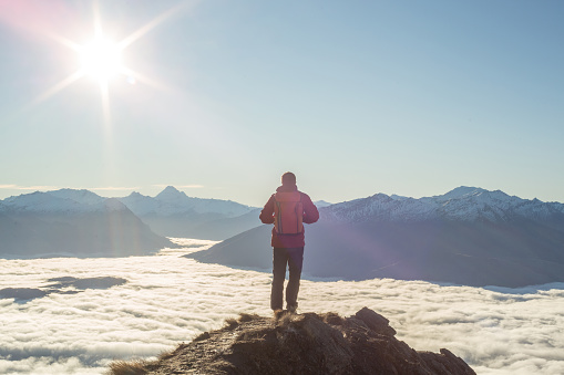 Young man hiker stands on a mountain peak above the clouds and contemplates the spectacular view, sun shining over the mountains. Shot in New Zealand on the South Island.
