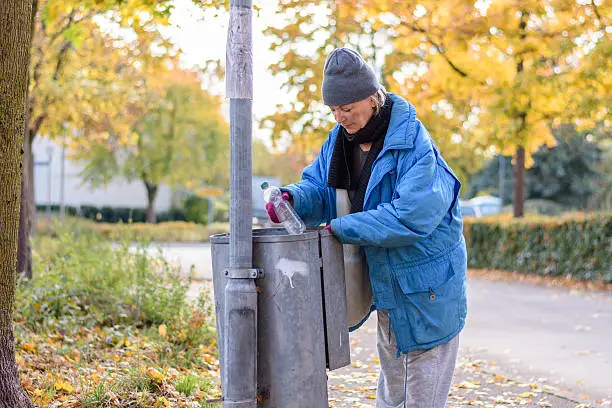 Indigent senior woman scrounging through a bin for food on an urban autumn street in a concept of homelessness and poverty
