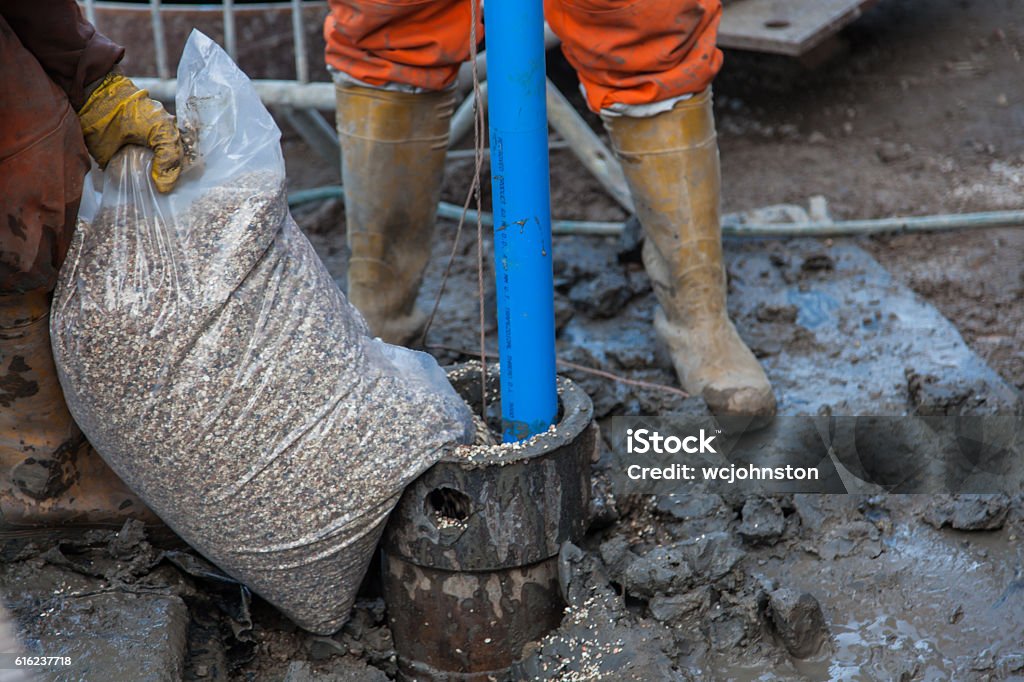 Men in orange overalls and boots work on a borehole Men in orange overalls and boots work on a borehole to fill the hole with gravel and a blue water pipe protrudes from the hole, the ground is muddy. Motion bluer on the gravel Filling Stock Photo