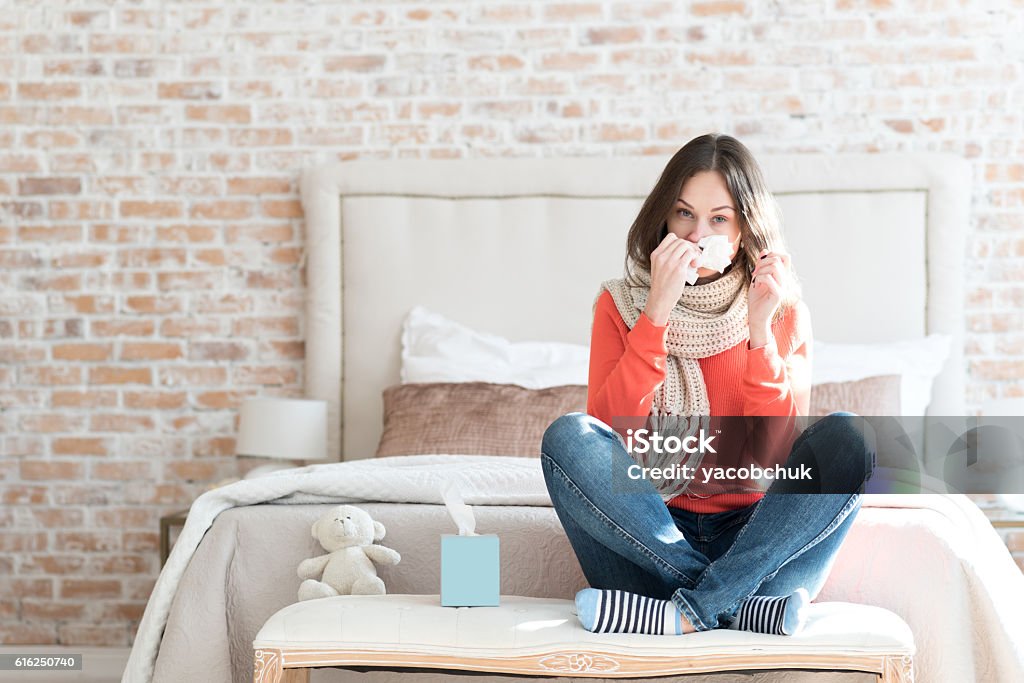 Pleasant gloomy woman suffering from cold Winter epidemics. Nice moody young woman sitting in a bedroom and wearing a scarf while suffering from cold Illness Stock Photo