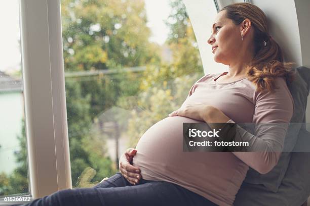 Mujer Embarazada Sosteniendo Su Vientre Mirando A Través De La Ventana Foto de stock y más banco de imágenes de Embarazada