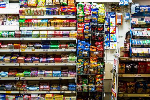 London, UK - October 15, 2016: a man working in a newsagent kiosk in Monument railway station in central London, UK. He is surrounded by the wares that he is selling, which mainly consists of colourful sweets and candy, and packets of crisps. The man's face is largely obsured, but he is of Indian ethnicity.