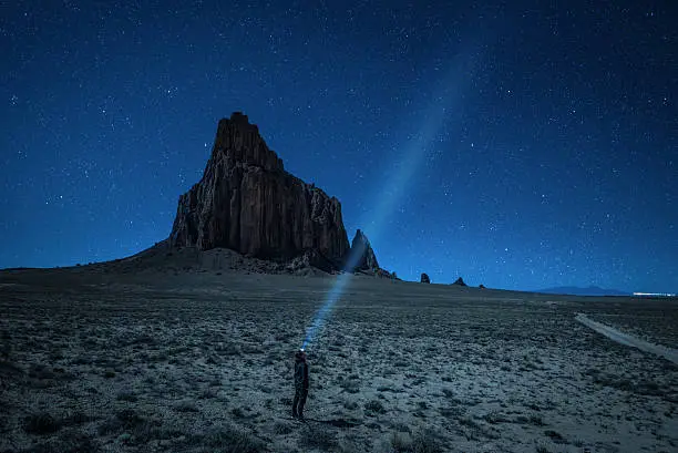 Hiker with a head lamp under the night sky with many stars near Shiprock. Shiprock is a great volcanic rock mountain rising high above the high-desert plain of the Navajo Nation in New Mexico, USA