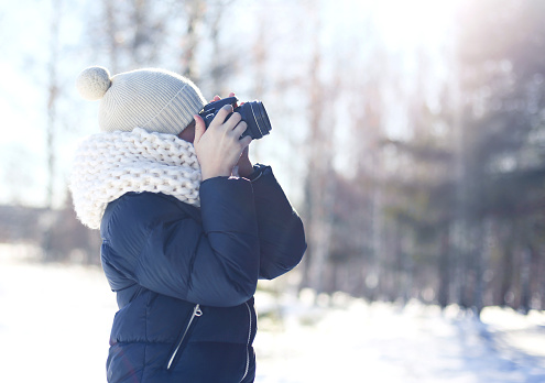 Child little boy photographer takes picture on the digital camera outdoors in winter sunny day over blurred forest background, view profile