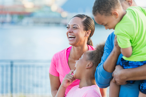 A happy African American woman laughing, standing outdoors with her young family. Her daughter is standing beside her, looking up affectionately. Her son and husband are out of focus in the foreground.