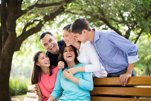 A latin family of five embracing and laughing together on a bench in a horizontal medium shot outdoors.
