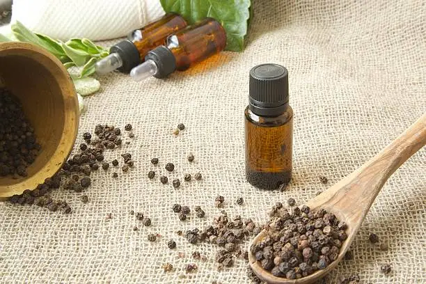 A dropper bottle of black pepper essential oil. Black pepper in the background.