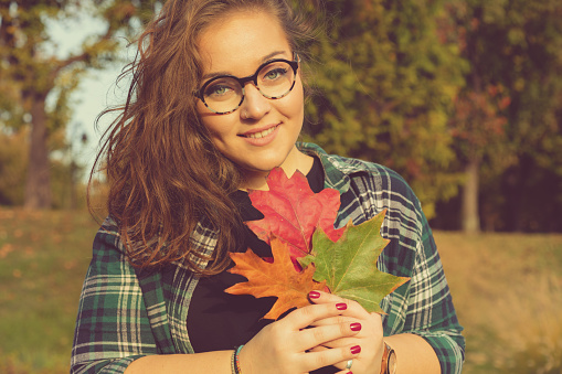 Smiling woman holding autumn leaves outdoors in colorful fall forest.