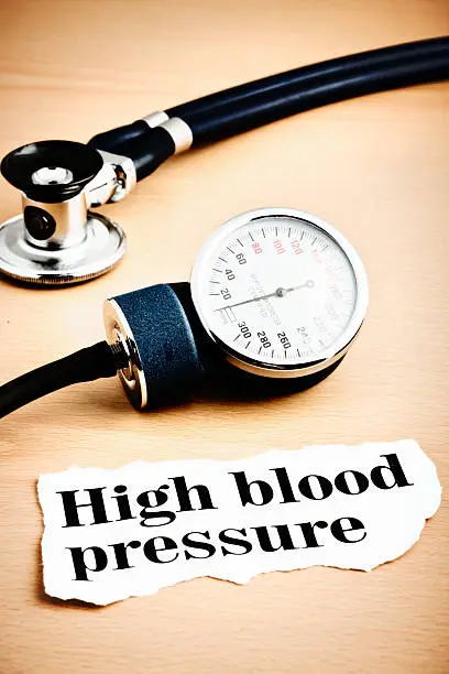 Printout reading 'high blood pressure' on a wooden desk with part of  a blood-pressure gauge and a stethoscope.