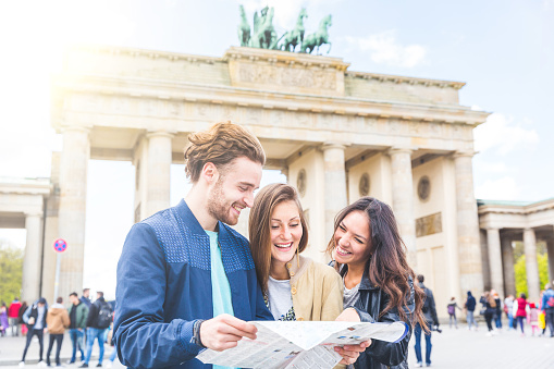 Multiracial group of friends visiting the city of Berlin. Two women and a man looking at a map with Brandenburg Gate on background. Lifestyle, friendship and tourism concepts with real people models.