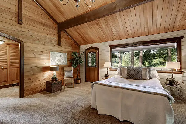 Wooden bedroom interior with high beamed ceiling, grey carpet floor and large bed with neatly arranged pillows.  Northwest, USA