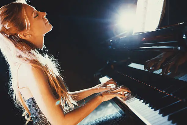 Closeup side view of a mid 30's female pianist playing at a concert. Her eyes are closed and she's enjoyng her performance. The stage is in complete dark except two spot lights on her.