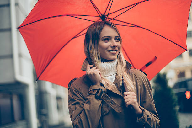 Attractive young smiling woman carrying umbrella and adjusting her coat while walking by the street