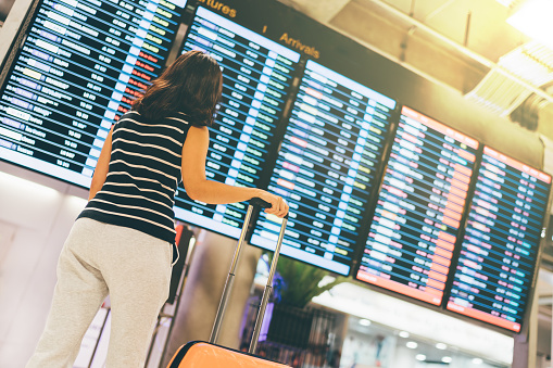 Asian woman traveler looking at flight information screen in an airport, holding suitcase, travel or time concept, warm light effect