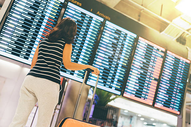 Asian woman traveler looking at flight information screen in airport Asian woman traveler looking at flight information screen in an airport, holding suitcase, travel or time concept, warm light effect delayed sign photos stock pictures, royalty-free photos & images
