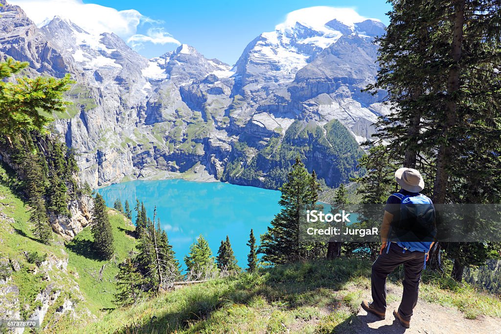 Hiking at Oeschinen Lake in Berner Oberland in Switzerland Male hiker is looking at turquoise Oeschinensee on the Heuberg - Oberbergli - Oeschinensee hiking trail at the UNESCO World Heritage Site Oeschinen Lake near Kandersteg in Berner Oberland region in Switzerland. The man is wearing hiking clothes, a sun hat and carrying a blue backpack. Switzerland Stock Photo