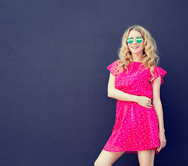 Summer Fashion Woman in Pink Dress at Dark Wall Fashion Woman in Pink Dress and Sunglasses at the Dark Textured Wall Background. Urban Fashion Girl in Summer Concept. Street Style Female. Toned Photo with Copyspace. dark pink dress stock pictures, royalty-free photos & images