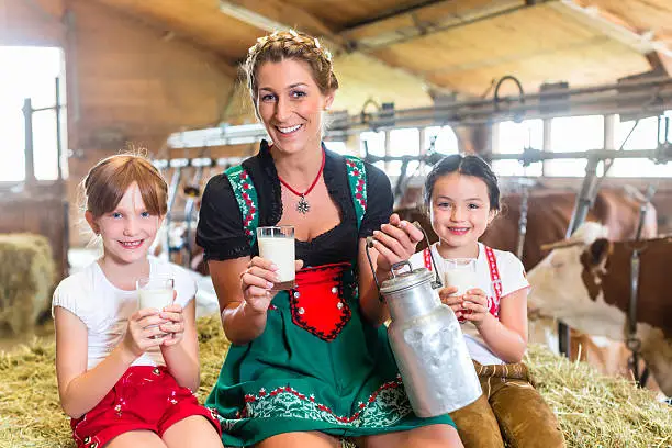 Bavarian mother sitting with children in cowhouse on hay bale drinking fresh milk