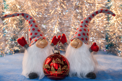 Two cheerful gnome with his hands up in red mittens and high caps holding red glass Christmas ball on silver background. Christmas or New Year picture.