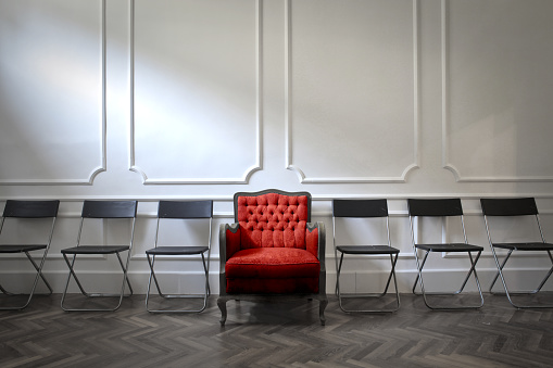 An exquisite red vintage arm-chair amid cheap black chairs standing in the row on the parquet floor in front of white wall in the classically designed room 