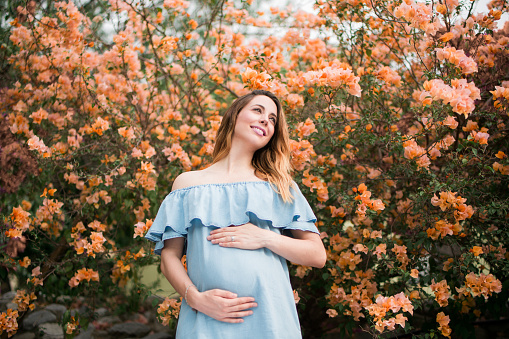 A cheerful pregnant woman holding her belly and smiling away in a horizontal medium shot outdoors with flowers in the background.