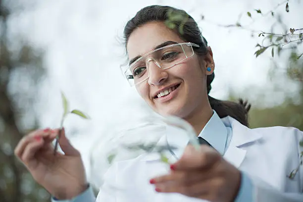 Outdoor low angle view of a happy young female botanist collecting sample of leafs in specimen holder. She is giving toothy smile while standing in natural environment. She is in her protective work wear and lab coat. One person, waist up, horizontal composition with copy space and selective focus.